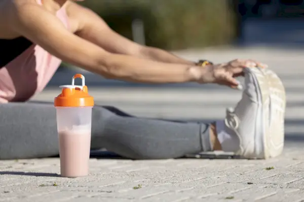 woman stretches on the cement of an outdoor park while wearing athletic clothing, with a smoothie drink next to her.