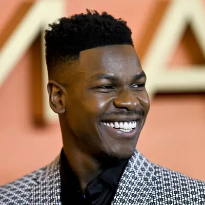 Close up of actor John Boyega with a fade haircut, against an out of focus orange background