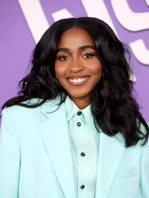 Ayo Edebiri smiling at camera in blue button up and blazer, with hair curled, middle part in butterfly cut