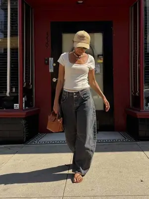 Person wearing nude ball cap, white tea, gray low rise barrel jeans from free people with brown bag and brown flip flops
