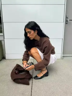 Woman wearing brown sweater, white skirt, black heeled Mary Janes, silver earrings, sunglasses, and brown suede handbag