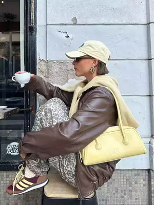 Woman wearing baseball cap, brown leather jacket, leopard print pants, brown and yellow sneakers, and butter yellow Marge Sherwood bag