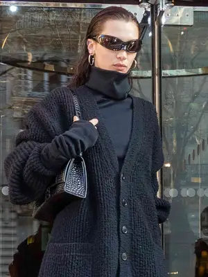 Bella Hadid wears a black turtleneck and cardigan, black leather shoulder bag, wraparound sunglasses, and silver hoop earrings
