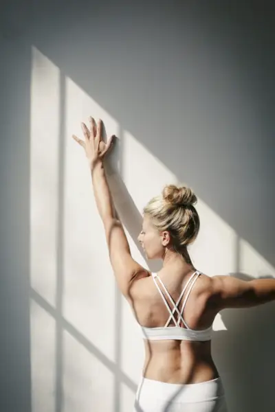 A blonde woman, in a white crop top and leggings, standing in front of a white wall, doing yoga, her arm raised, touching the wall.