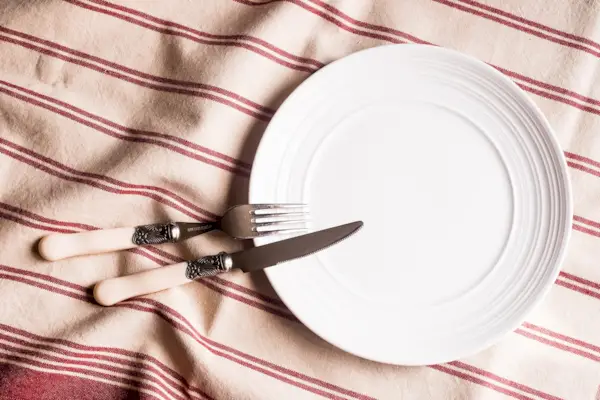 Empty white plate, fork and knife on a red and white linen striped napkin, top view. Image with copy space. Kitchen table with a towel and a plate - top view with copy space.