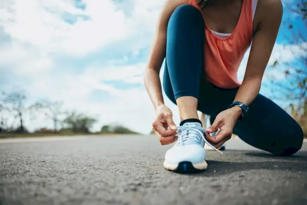A young woman runner is outside in the morning, preparing for a jog. She is seen bending down, tying her shoelaces, ensuring her running shoes are securely fastened before she begins her run