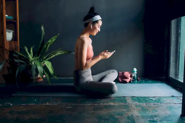 Choosing Workout Music: Asian Woman With Earphones Sitting on the Exercise Mat and Setting Up a Playlist on Her Smarphone