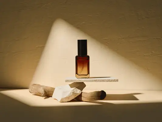 A composed image of a bottle of perfume resting above a piece of wood and ceramic