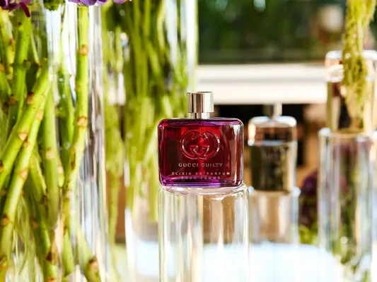 Gucci Guilty Elixir perfume bottle with light purple color atop glass cylinder and surrounded by greenery