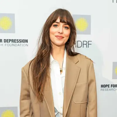 Dakota Johnson with loose waves and wispy bangs wearing a beige blazer