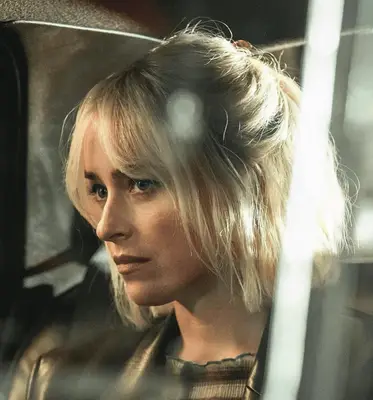 Close up of Dakota Johnson in a black shirt, standing against a white wall, her hair long and dark with choppy bangs