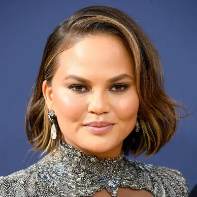 Chrissy Teigen attending an event, smiling with short styled hair and wearing statement jewelry