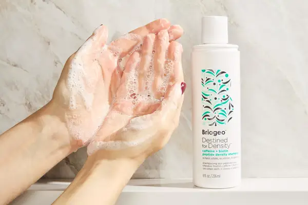 A person lathering Briogeo Destined For Density Caffeine + Biotin Density Shampoo in their hands next to the bottle
