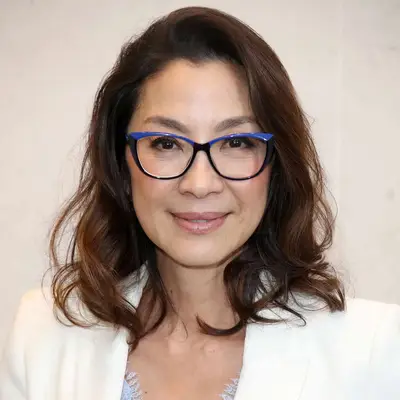 Michelle Yeoh wears a shoulder-length wavy hairstyle and blue and black glasses