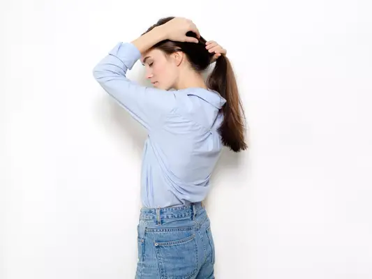 A woman in a button down blue shirt and jeans, pulling her hair back into a ponytail with her other hand on her head