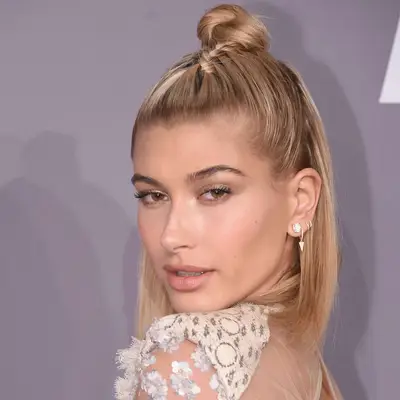 Hailey Bieber looking over shoulder at camera, zoomed in on face with short blonde straight bob and top section pulled back tightly with braid going back into a twisted bun on top of head