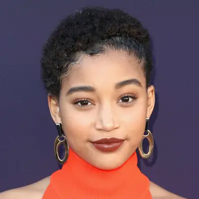 Amandla Stenberg attends an event with short, natural afro and smooth edges, a high orange turtleneck and bold deco gold earrings