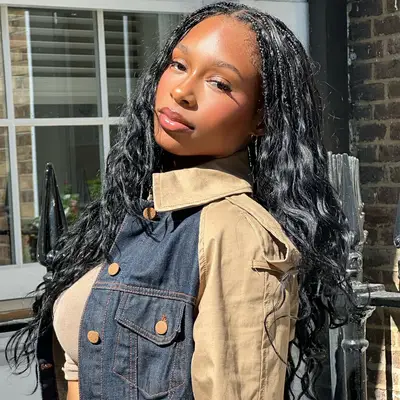 A woman with long braided hair, wearing a denim and beige jacket, standing outdoors with a casual expression