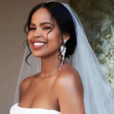 Sabrina Elba wears a veil and low updo with loose curls and statement earrings