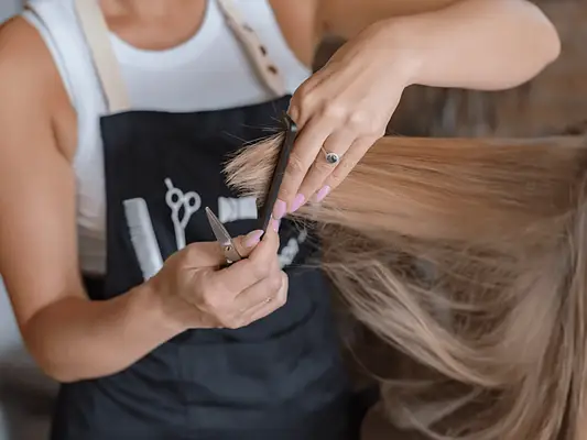 A woman in an apron holding a comb and scissors, cutting the ends off of another woman