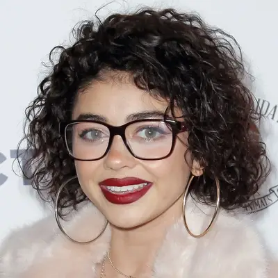 Sarah Hyland wears a layered curly bob hairstyle, pale pink fur coat, hoop earrings, red lipstick, and glasses at the 2018 Winter Wonderland Toys for Tots Party in Los Angeles