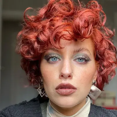 Beauty creator wears short red curly hair, brushed brows, and blue-green eyeshadow