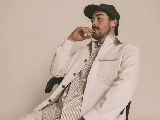 A man with stubble and a mustache wearing a baseball cap, button down shirt and jacket