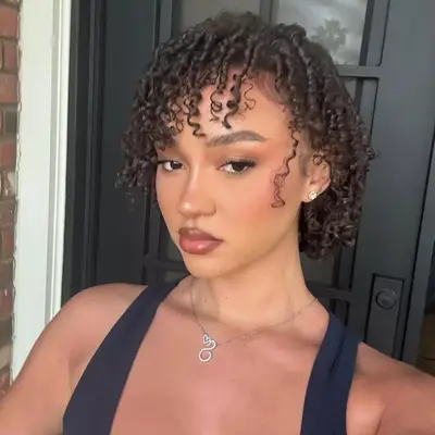 A person with short curly hair looking at the camera, wearing a necklace