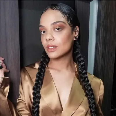 Tessa Thompson wears braided pigtails, a warm-toned makeup look, and a gold satin blazer