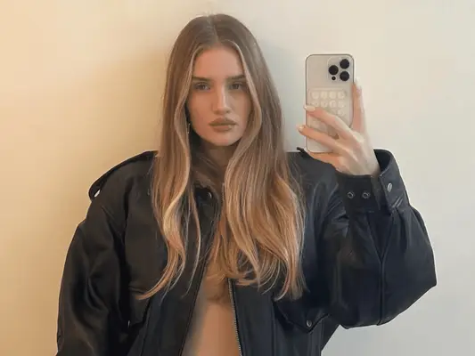 Rosie Huntington-Whiteley holding her cell phone and taking a mirror selfie of her glossy blonde hair with brightened money pieces