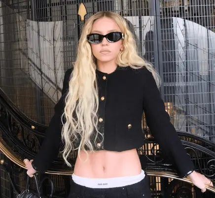Sydney Sweeney poses out front of the Miu Miu show in Paris with long, blond wavy hair wearing a cropped blazer and sunglasses