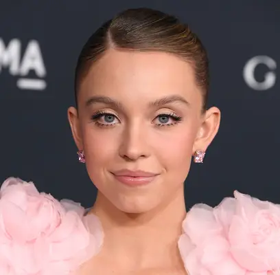 Sydney Sweeney at a LACMA event in a fluffy pink sleeved dress, pink studded earrings and soft pink tinted makeup with a sleek side parted updo. 