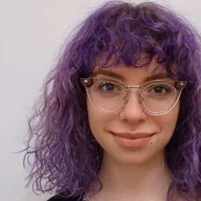 Woman with curly purple hair with bangs and cat-eye glasses