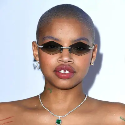 Slick Woods wears dyed lavender buzzcut and eyebrows and hexagon-shaped sunglasses