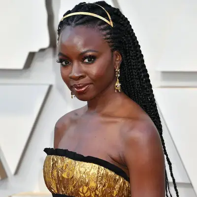 Actor Danai Gurira on the red carpet with a half cornrow, half box braid hairstyle, her hair and ears adorned with gold accessories