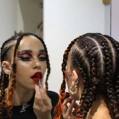 FKA Twigs applying red lipstick, viewed from the back, her cornrows in view