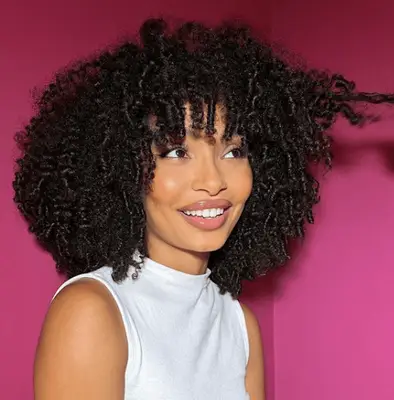 yara shahidi with coily curly hair and bangs in front of fuschia background