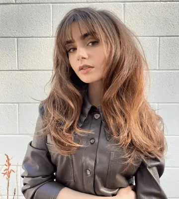 lily collins with wispy bangs and long blown out hair in leather shirt against white brick wall