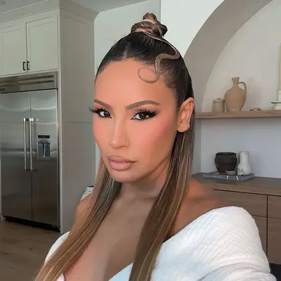 Person with long straight hair styled in a top knot, wearing an off-shoulder garment, standing in a modern interior