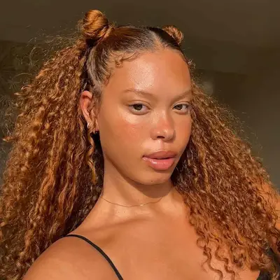 A person with curly hair styled in two buns and loose curls looks at the camera