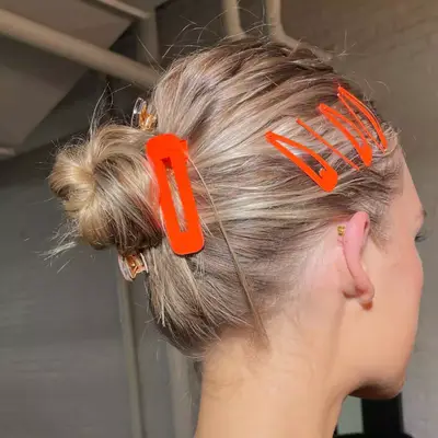 model with blonde hair pulled back into bun, side view, with bright orange barrettes pinning hair back on the side, and one giant orange hair clip by the side of the bun