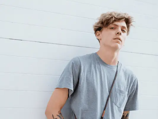 A man with flowing side swept hair standing against a wall