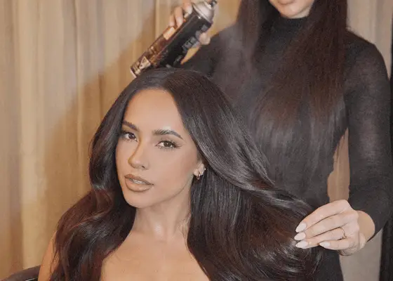 A woman sitting in a chair while a hairstylist sprays her long brown hair with hairspray