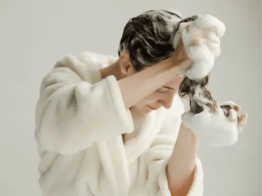 A woman in a bathrobe with wet hair and shampoo suds on her hands and hair