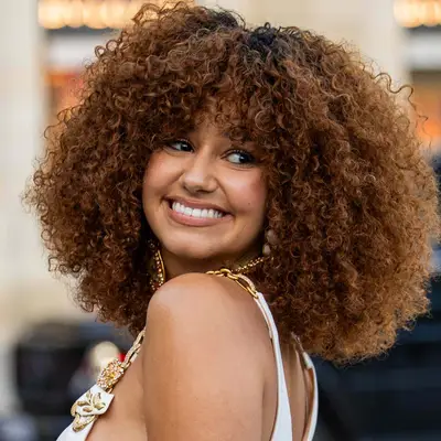 Lena Mahfouf looks over her shoulder with her natural coily curls coated in a golden brown color