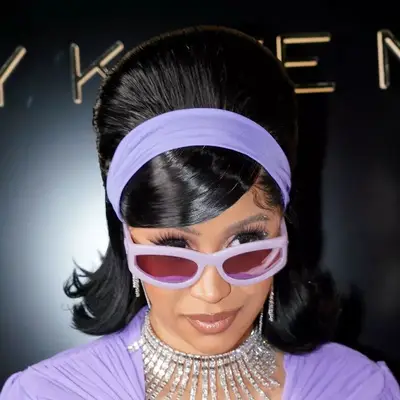 Cardi B sporting a bouffant hairstyle with a purple headband and matching sunglasses, along with a purple outfit and diamond jewelry at 2021 Paris Fashion Week.