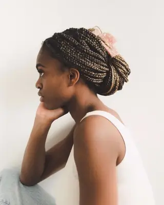 A person sitting in profile view, resting their chin on their hand with braided hair clipped back and wearing a tank top
