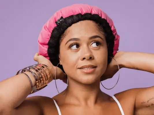 Model in front of a purple background with silver bracelets and hoop earrings wearing a pink heat cap.