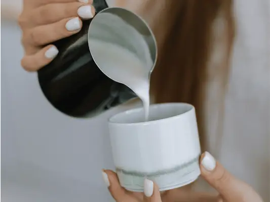 A woman holding frothed milk in a silver frothing pitcher and pouring it into a cup