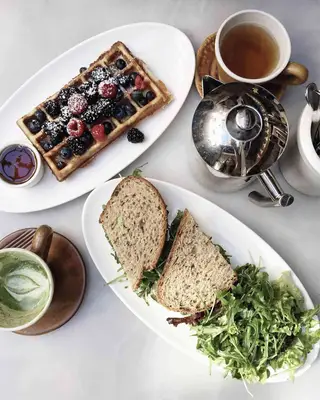 Table top with waffles and berries; sandwich halves with salad; matcha cup; tea cup; and tea kettle